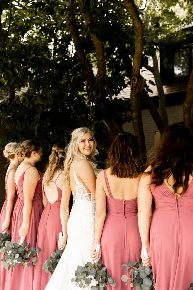 Bride and bridesmaids stand shoulder to shoulder facing away from photo, showing off the gorgeous backs of their dresses, as bride turns to flash a smile at the photographer.
