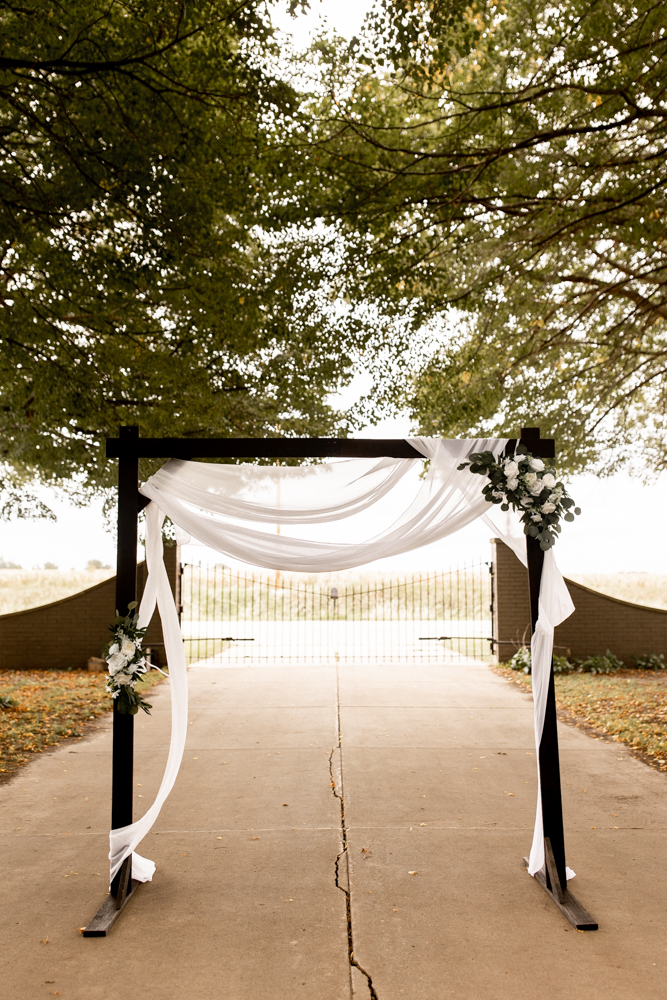 Beautiful wedding arch with hanging white drapery and subtle white with greenery floral arrangements on each side. 