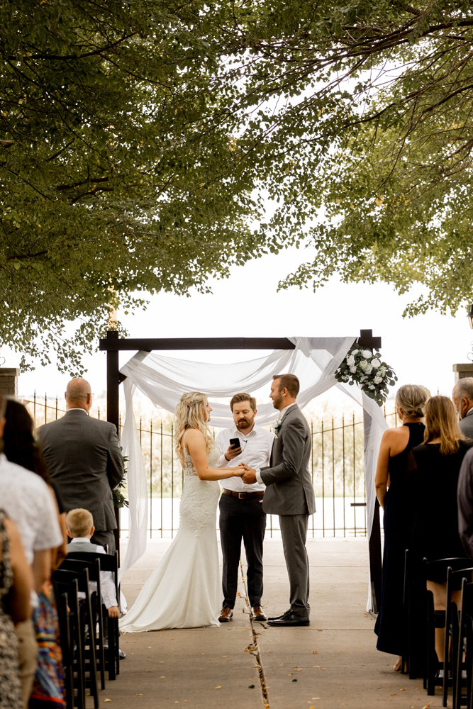 Bride and groom stand holding hands at the altar of their gorgeous wedding with beautiful outdoor backdrop. 