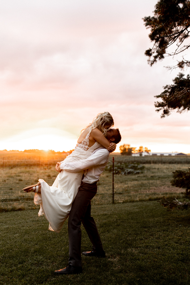 Husband lifts his wife and kisses her as the sun is setting for a picture perfect moment in the background. 