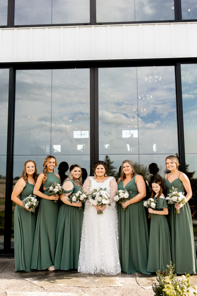 Bride and her bridesmaids in their beautiful sage green dresses pose in front of Little Lights on the Lane Wedding Venue. 