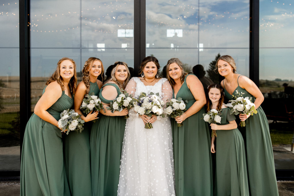 Bride and her bridesmaids in their beautiful sage green dresses pose in front of Little Lights on the Lane Wedding Venue. 