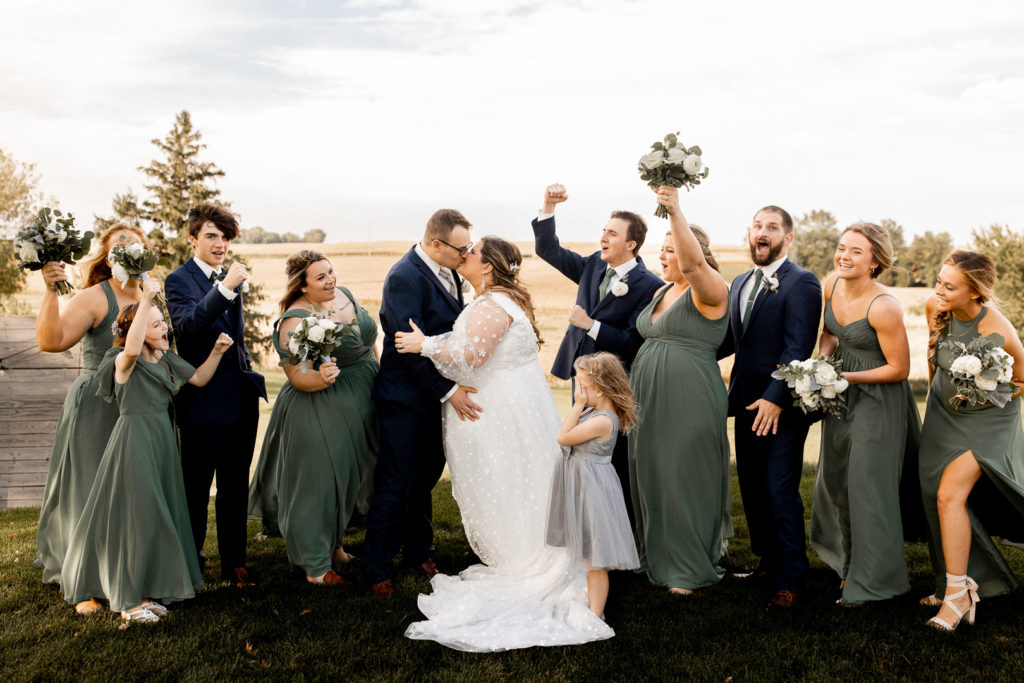 Bridesmaids and groomsmen all cheer and throw their hands in the air as the bride and groom kiss. Beautiful flowers in the air and bridal party having a fun time! 