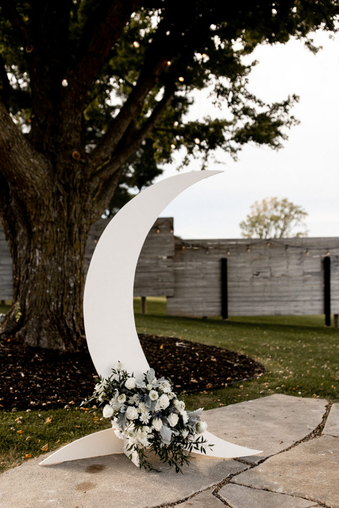 Crescent Moon and beautiful floral arrangement displayed at the bottom of the moon serve as the decor at the end of the wedding aisle. Details of decor and wedding day captured by Iowa Wedding and Lifestyle Photographer. 