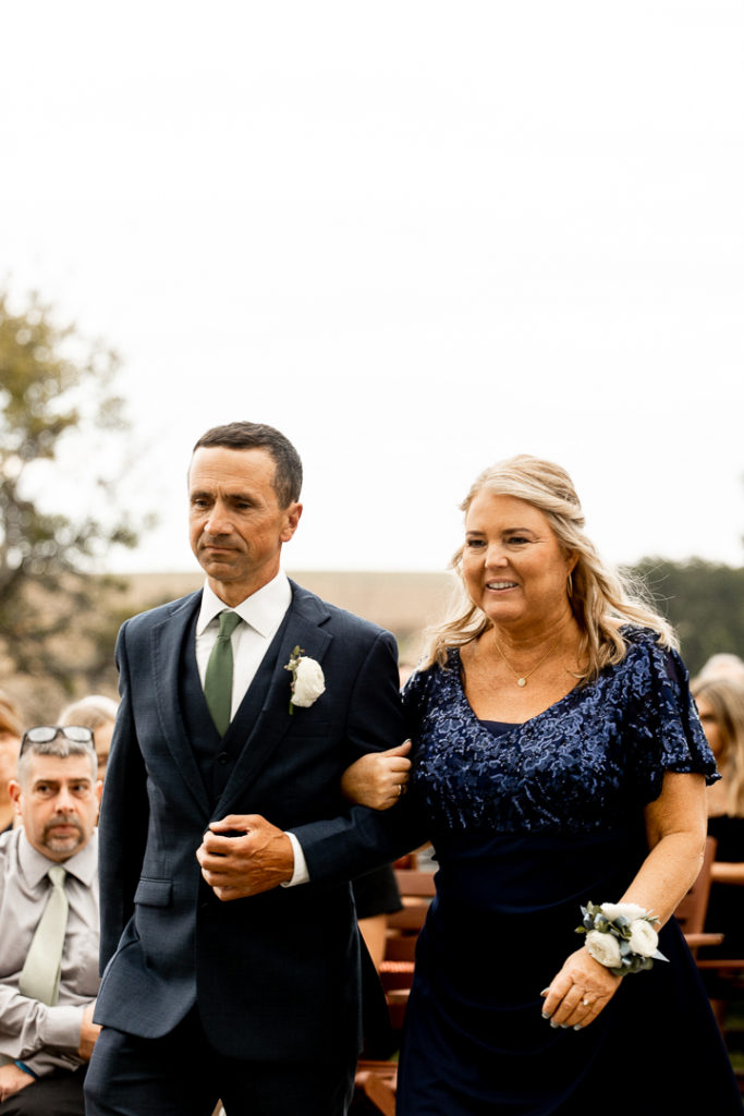 Groom's mom and dad walk down the aisle to be seated in reserved seating at the front of the wedding ceremony to witness their son marry his bride. 