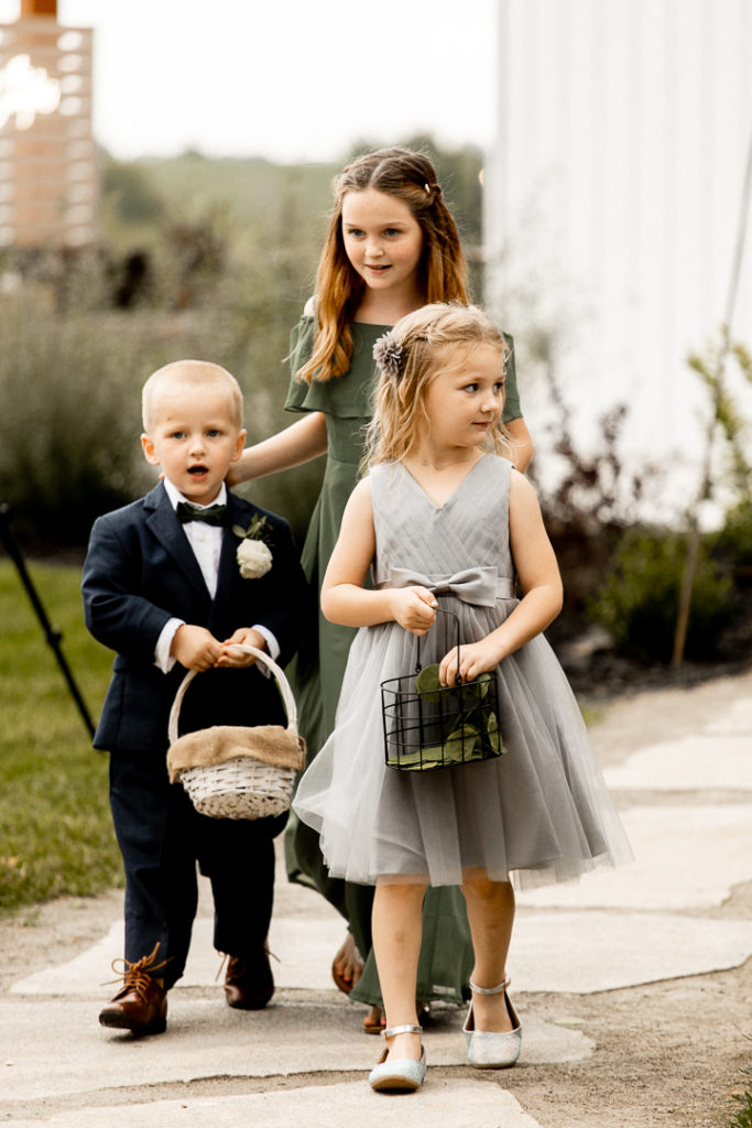 Flower Girls and Ring Bearer walk down the aisle throwing flowers and carrying the rings just moments before the bride herself makes her way down the aisle at her gorgeous wedding ceremony at Little Lights on the Lane in West Branch, Iowa. 