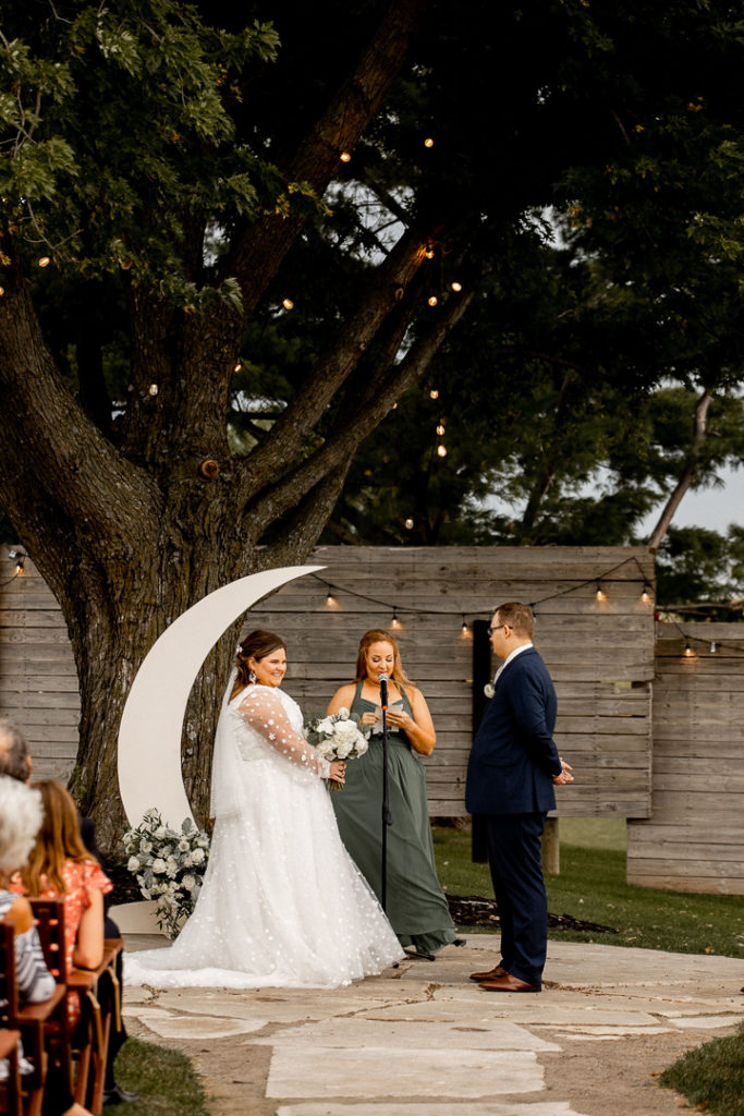 Bridesmaid helps with a reading for the wedding ceremony. Moon decor behind her and lights hanging from the tree offer stunning backgrounds. 