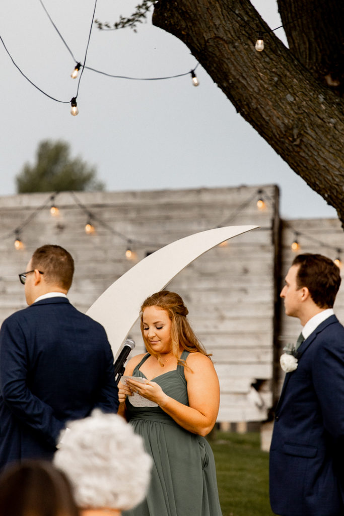 Bridesmaid helps with a reading for the wedding ceremony. Moon decor behind her and lights hanging from the tree offer stunning backgrounds.