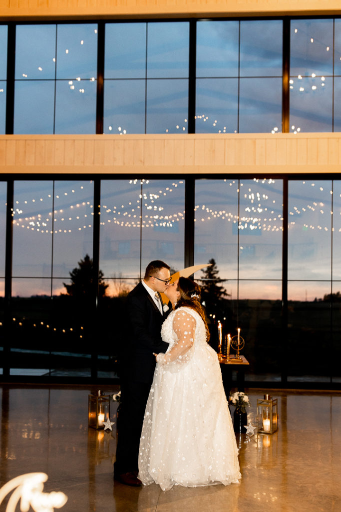 Bride and groom share their first dance as husband and wife and end it with a kiss. Lights reflect off the glass of the venue, creating the most stunning backdrop. 