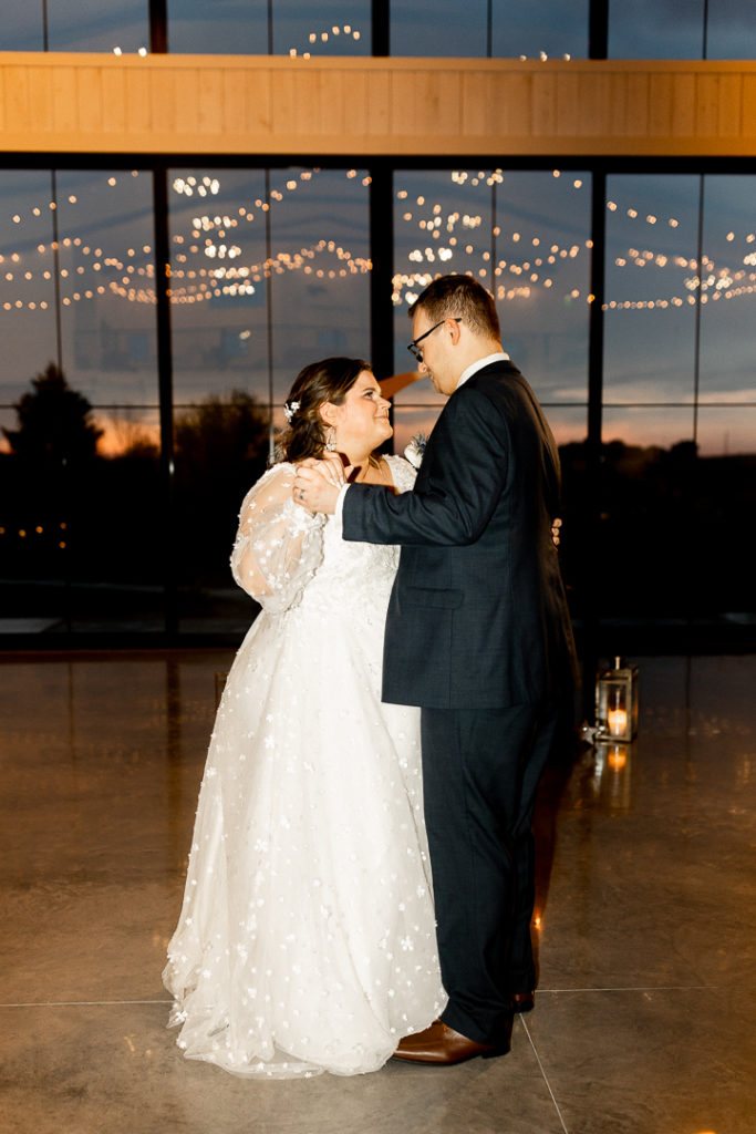Bride and groom share their first dance as husband and wife. Lights reflect off the glass of the venue, creating the most stunning backdrop. 