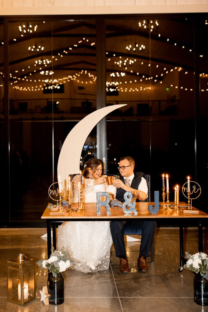 Husband and wife clink their drinks in a cheers at the head table. Head table complete with moon and stars decoration and wedding venue is beautifully decorated and photographed by Midwest Wedding Photographer. 