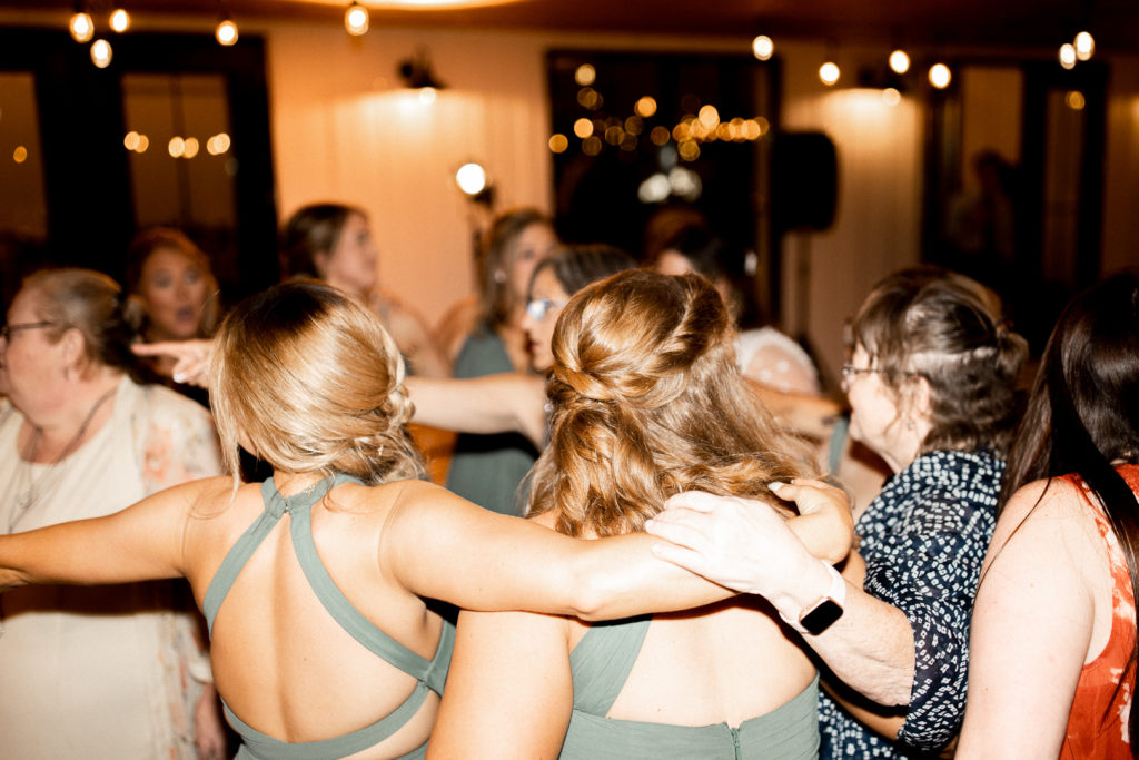 Hands over shoulders create a big circle on the dance floor as friends and family of the bride and groom come together to dance and celebrate the couple. Fun moments of the reception and wedding day captured by Iowa Wedding and Lifestyle Photographer. 