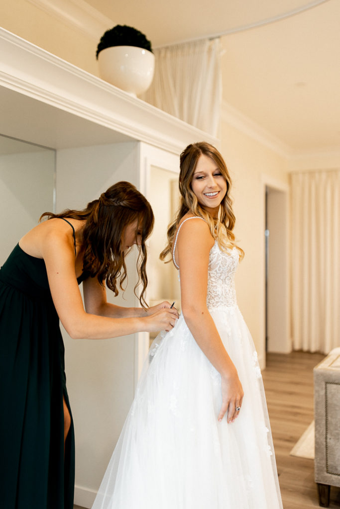 Bride peers over her shoulder and smiles for the photographer as bridesmaid continues with buttoning up the bride's wedding dress. 
