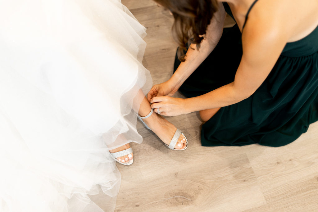 Sweet photo capturing special moments of the wedding day as a bridesmaid helps the bride fasten her shoes, putting the last touches on her stunning wedding day look.