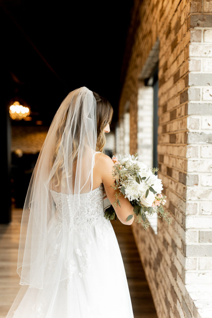 Bride facing away from the camera to show off the stunning back of her wedding dress and her gorgeous flowing veil.  Bridal bouquet hangs over her arm and gorgeous Iowa wedding venue offers nice background. 
