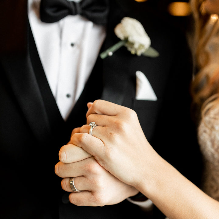 Close up detail photo of bride and groom holding hands and showing off their wedding rings. Hands placed in front of groom's chest showing the little details of grooms attire. 
