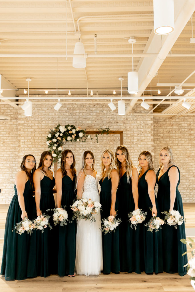 Bride and bridesmaids pose for wedding photo with their bouquets down at their side. Stunning royal green bridesmaid dresses. Wedding arch with amazing florals shown in the background of the photo at the Iowa Wedding Venue Toast. 
