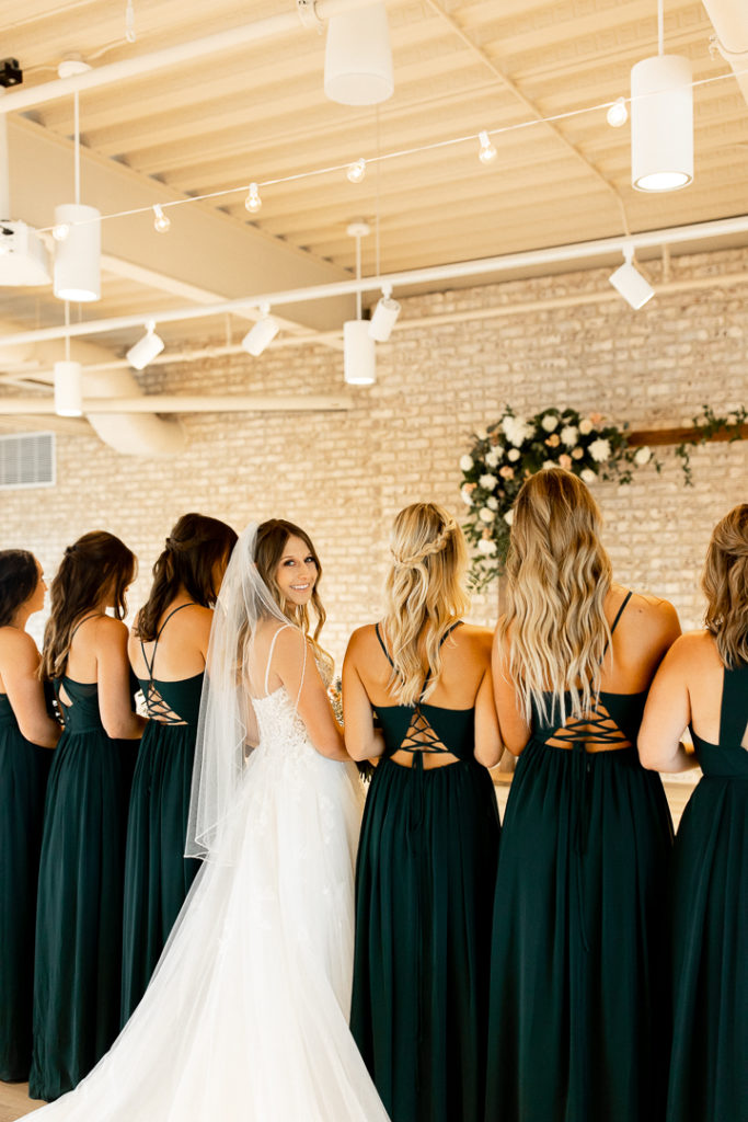 Bridesmaids and bride all face away from the photographer to show the back of their dresses, but bride peers over her shoulder and shows a smile. 