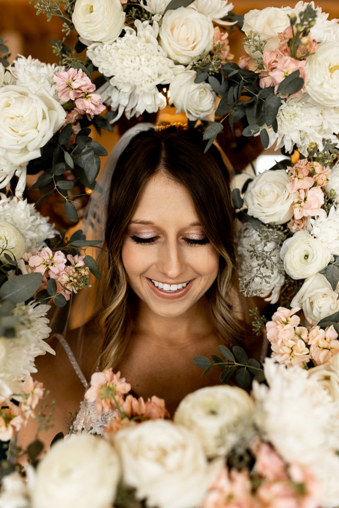 Bride peeks her head through a frame of stunning wedding flowers for a photo. Bride looks down to showcase her beautiful eye makeup. 