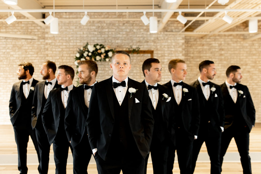 Groom in the center with groomsmen posing for the photographer behind him in a V shape. Groom looks at the camera as the groomsmen all look outward. 