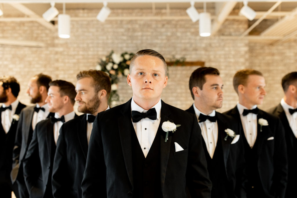 Zoomed in photo of the groom with his groomsmen posing for the photo behind him. Again, groom looks at the camera while groomsmen face outward. 