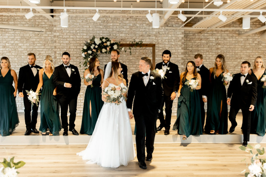 Bride and groom look at each other smiling as their wedding party walks and follows closely behind. Gorgeous backdrop with the wedding arch and Iowa Venue Toast.