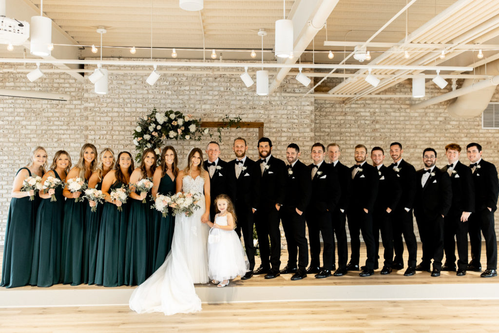 Bridesmaids and groomsmen line up on the sides of bride and groom all standing in a line posing for the Iowa Wedding Photographer. Beautiful wedding arch and floral bouquets in the background. 