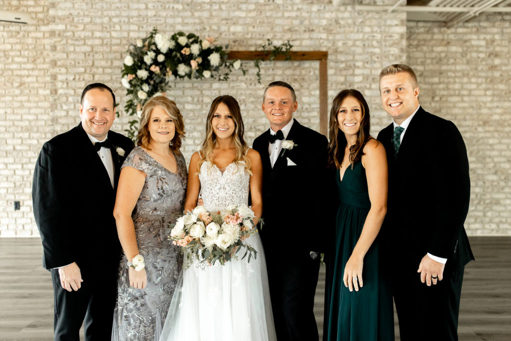 Sweet family photo with mom, dad, and siblings. Bride and groom smile and pose for the photo in the middle of the family 