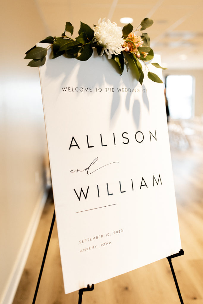 Beautiful white sign with sharp black writing to welcome their guests to their wedding ceremony. Flowers and greenery drape from the top of the sign. 