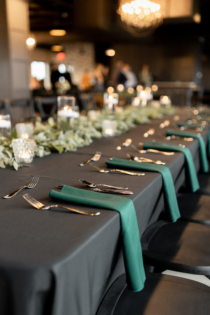 Beautiful details captured before guests arrive at the gorgeous Iowa Wedding Venue - Toast - in Ankeny, Iowa. Black table cloths with royal green napkins and tablewear are displayed. Stunning florals run down the center of the table to create gorgeous display for tablescapes. 
