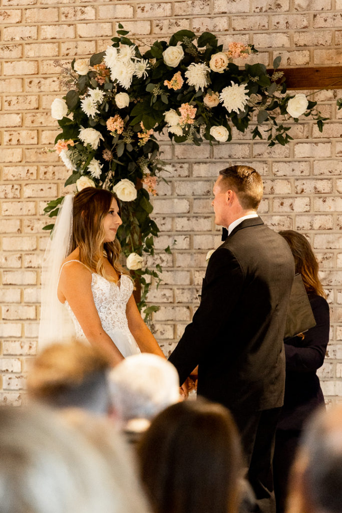Bride and groom hold hands and look into each other's eyes during their wedding ceremony, careful to take in every moment. Beautiful wedding arch with stunning florals draping over in the background. 
