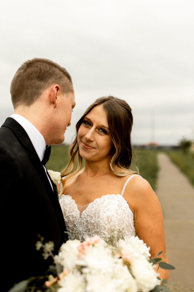 Husband and wife face each other as husband smiles adoringly down at his bride and she looks at the camera and offers soft smile. Beautiful bridal bouquet peeking up from the bottom of the photo. Gorgeous outdoor Iowa Wedding photos captured by Midwest Wedding Photographer. 