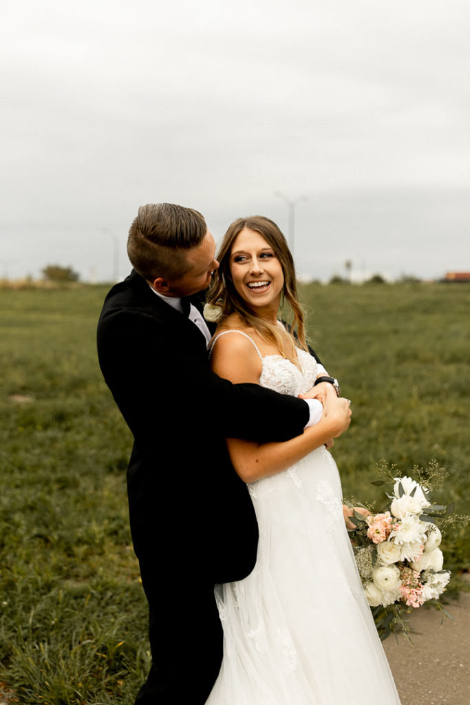 Husband and wife pose for the photographer with husband's arm wrapped around wife and smiling at her as she looks over her shoulder at him and smiles for the wedding photographer. Gorgeous green grasses in outdoor Iowa Wedding photos. 