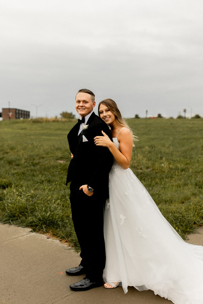Groom stands proudly in front of his bride as she leans in behind him, wrapping an arm around his. Beautiful outdoor wedding photos in the Iowa fall. 