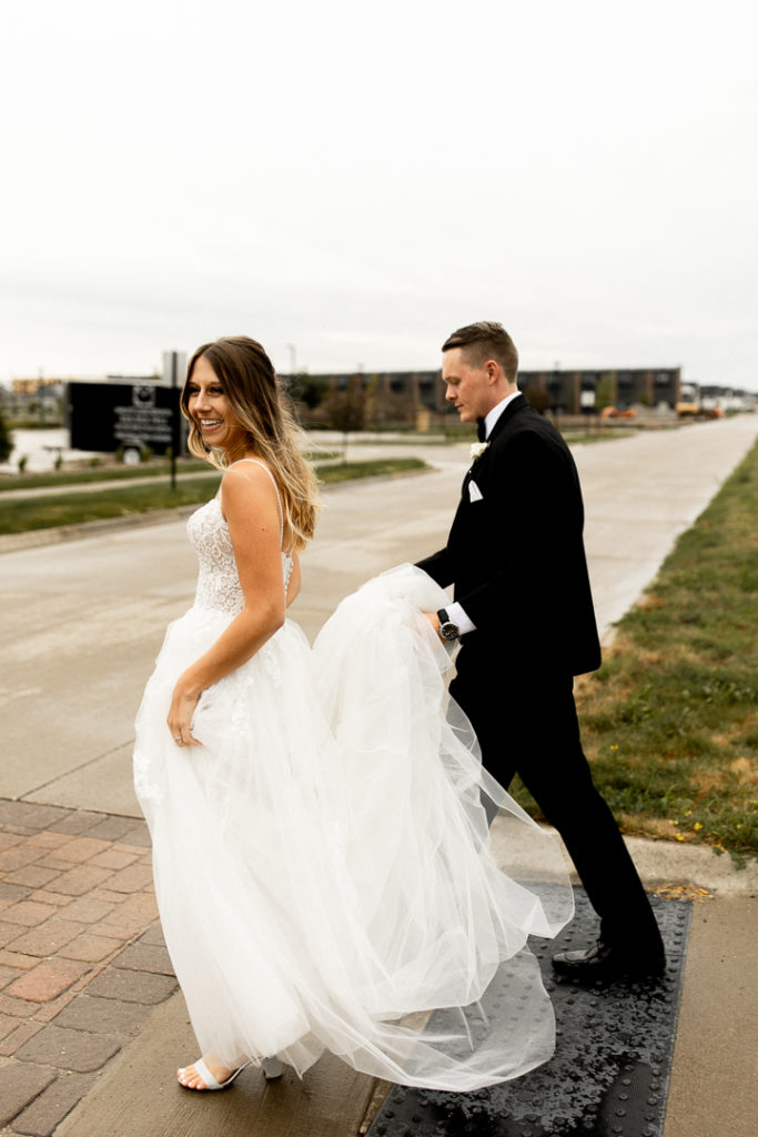 Groom carries his bride's wedding dress train as they cross the street. Rainy day in Iowa makes for stunning wedding photos. 