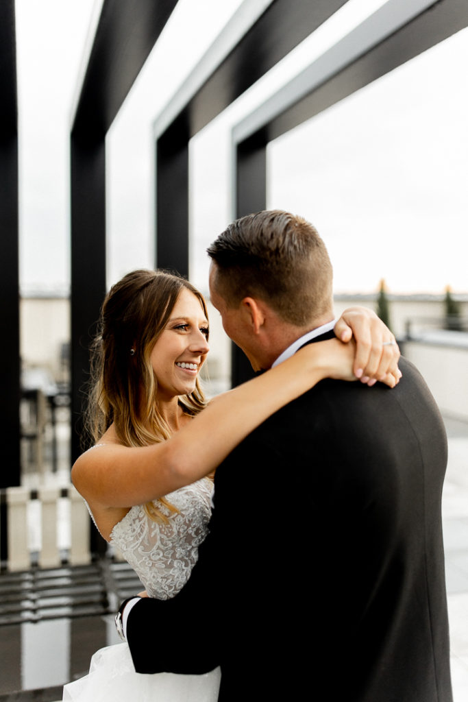 More photos on black patio with tall structures overhead. Bride and groom wrap their arms around each other's necks and stare lovingly into each others eyes as they flash a smile. 