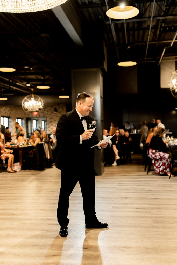 Dad of the bride begins with his speech, as the bride and groom watch and listen with smiling faces. Iowa Wedding Event Venue offers beautiful decorations and backdrops all around for these photos. 