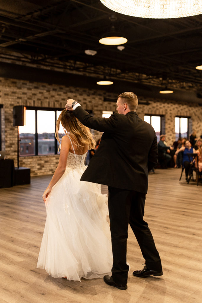 Bride and groom share their first dance as husband and wife with beautiful light fixture hanging above them at stunning Iowa Wedding Venue Toast in Ankeny Iowa. Husband spins his wife during dance. Sweet moments and memories captured by Iowa and Midwest Wedding Photographer. 