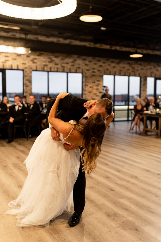 Bride and groom share their first dance as husband and wife with beautiful light fixture hanging above them at stunning Iowa Wedding Venue Toast in Ankeny Iowa. Husband dips his wife and kisses her during dance. Sweet moments and memories captured by Iowa and Midwest Wedding Photographer. 