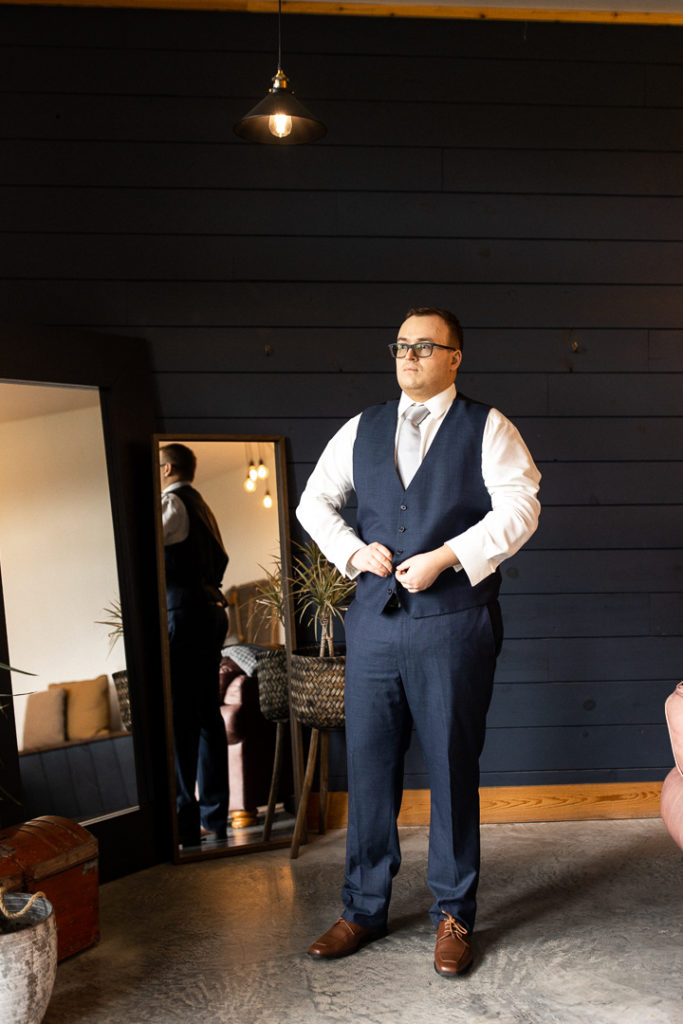 Groom preps for his wedding day as he buttons up his suit vest. 