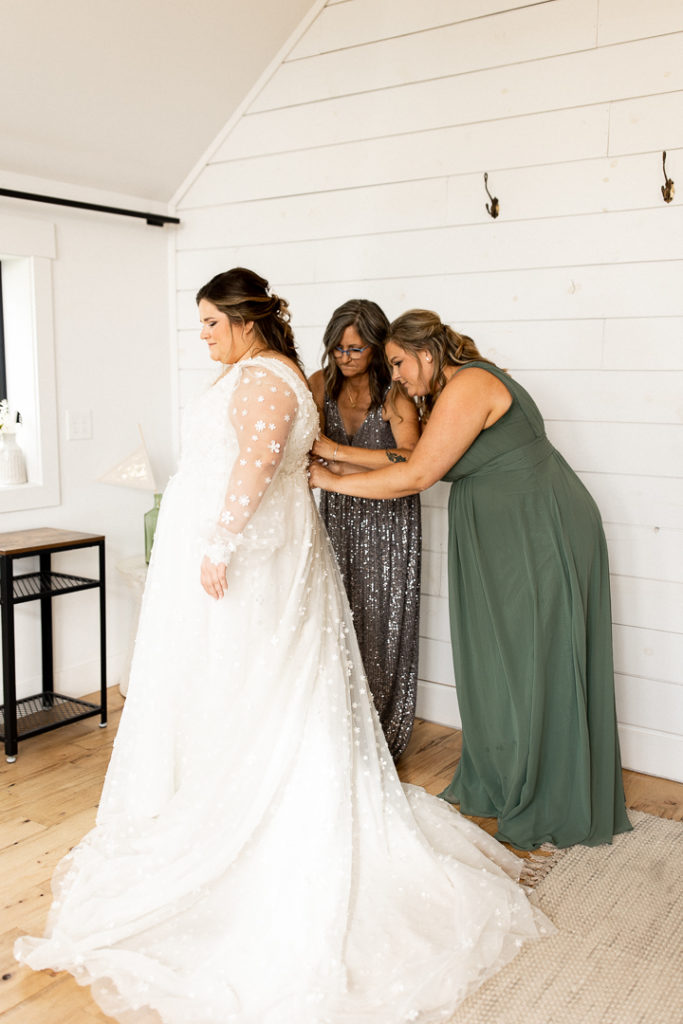 Bride's mom and bridesmaid help the bride put the finishing details on her wedding day look as they button her stunning long sleeve dress. 