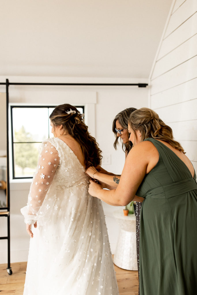 Bride's mom and bridesmaid help the bride put the finishing details on her wedding day look as they button her stunning long sleeve dress. Special memories shared with family captured by Iowa Wedding Photographer. 