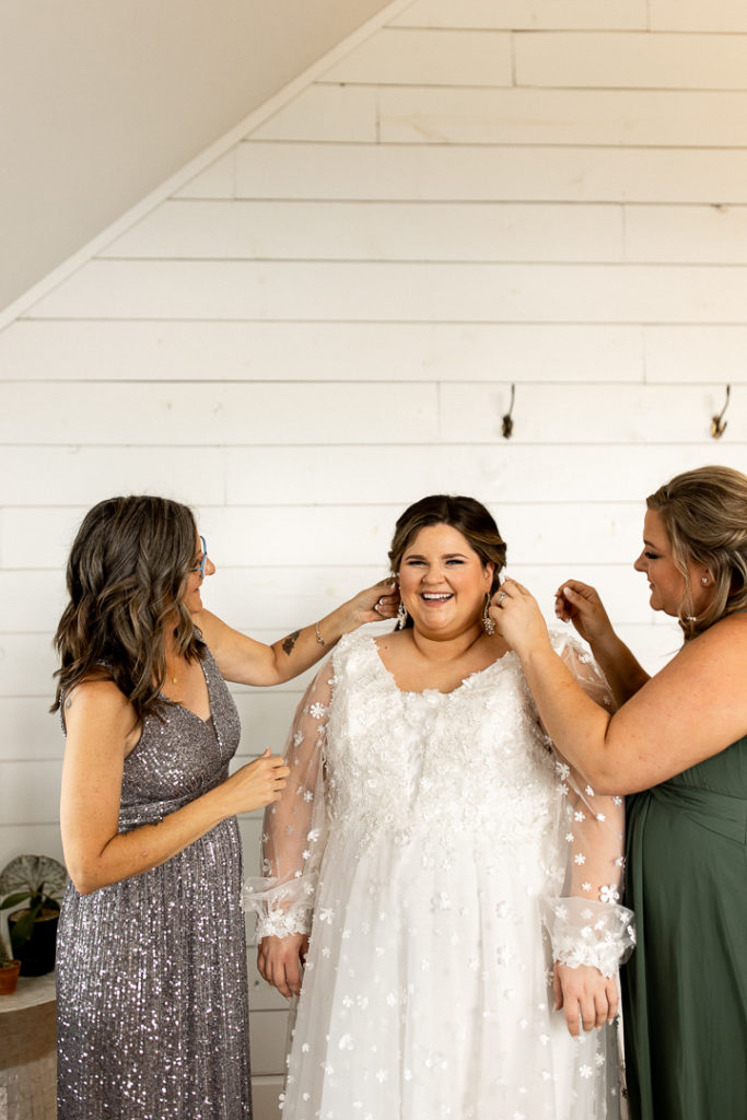Bride flashes huge smile for the camera as her mom and a bridesmaid helps her to put the finishing touches on her wedding day look. Sweet memories to treasure forever captured by Midwest Wedding and Lifestyle Photographer. 