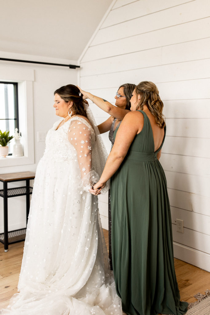 Bride's mom and bridesmaid help the bride put her veil in her beautiful bridal hair. Special family moments as the bride holds hand of bridesmaid. 