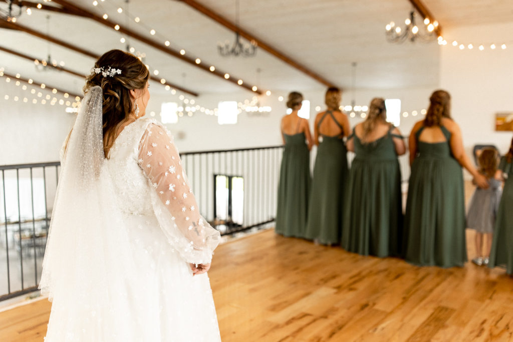 Bride stands behind bridesmaids as they are about to see her for the first time in her dress and completely ready for her wedding day. 